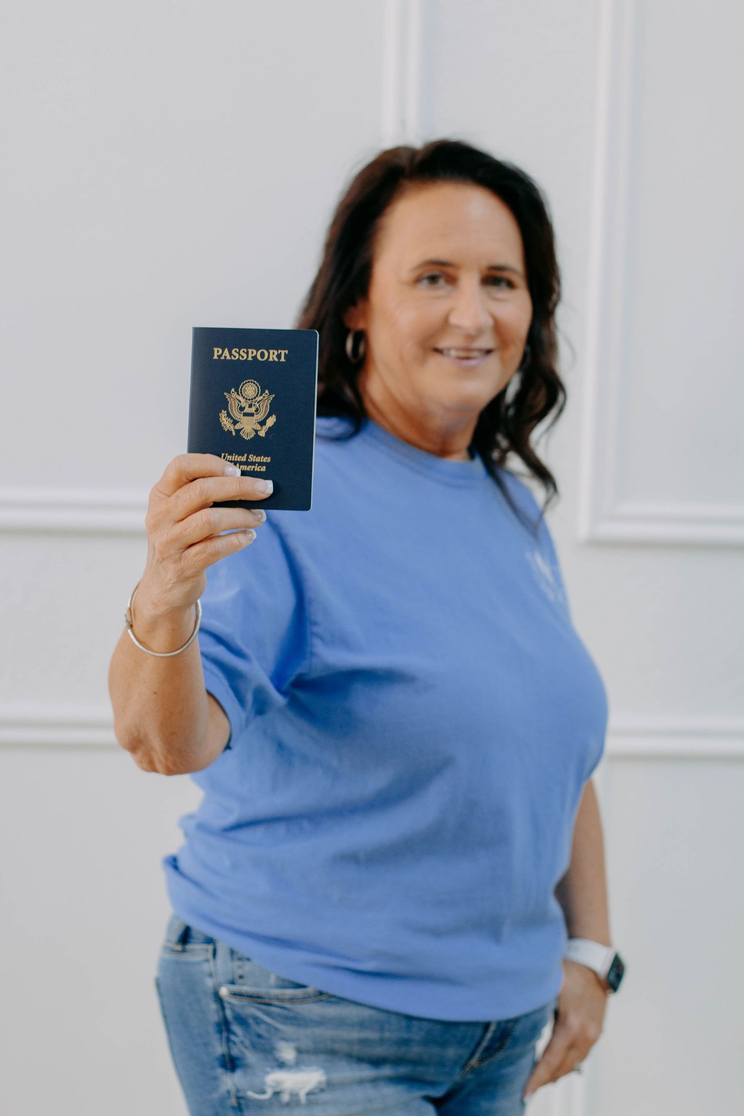 Woman in blue t-shirt smiling while holding a US passport toward camera.