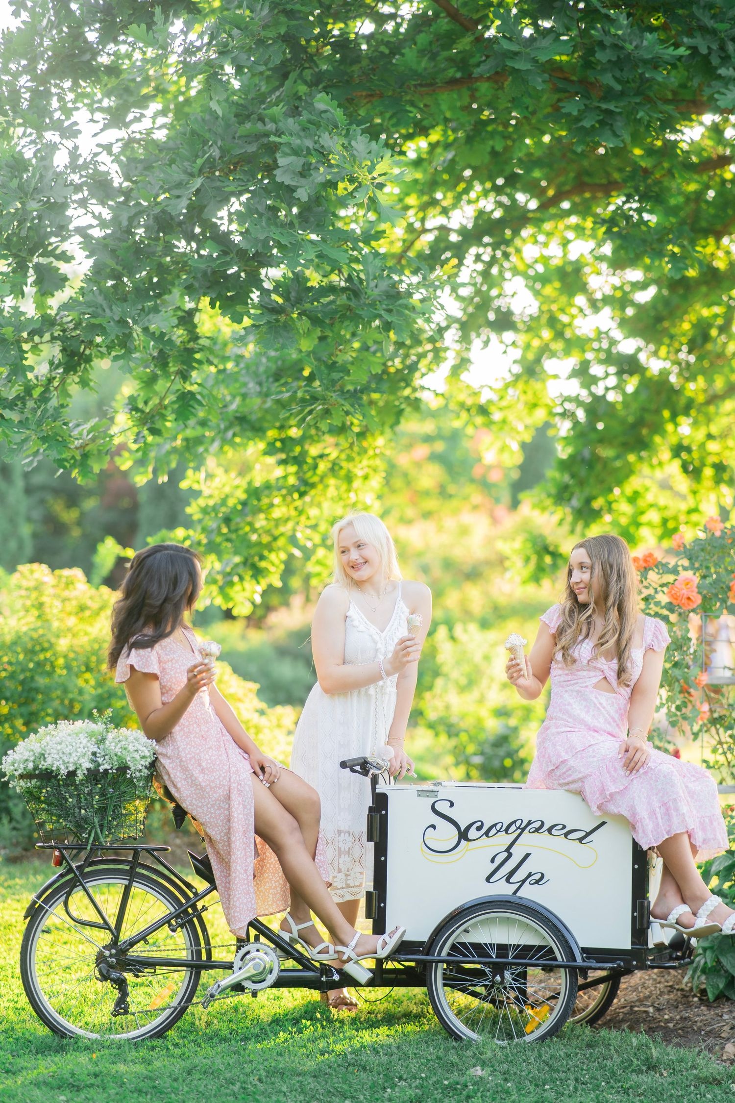 Three women in pink dresses gathered around a Scooped Up ice cream cargo bicycle in park.
