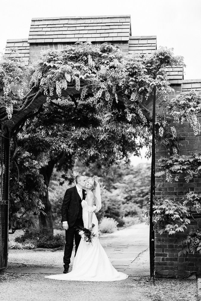 Black and white photo of a bride and groom kissing under a wisteria-covered archway at an elegant garden wedding.