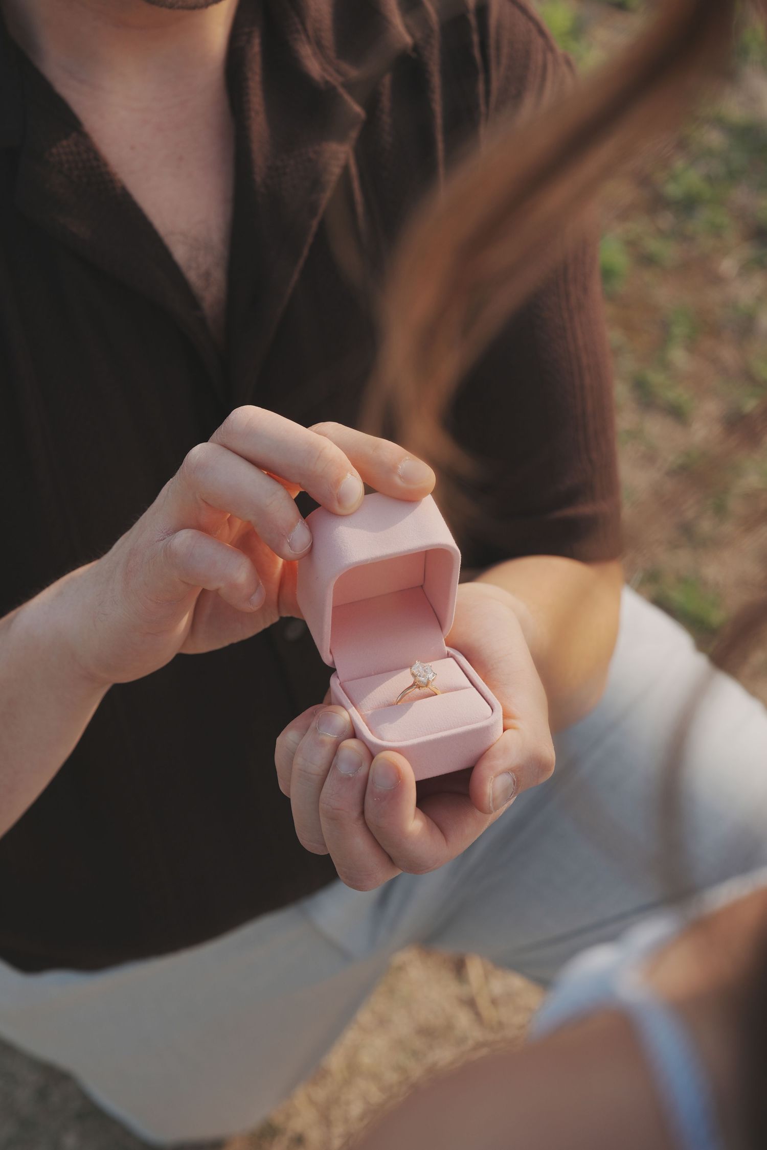Person opening a pink velvet ring box with a diamond engagement ring inside during a romantic outdoor proposal.
