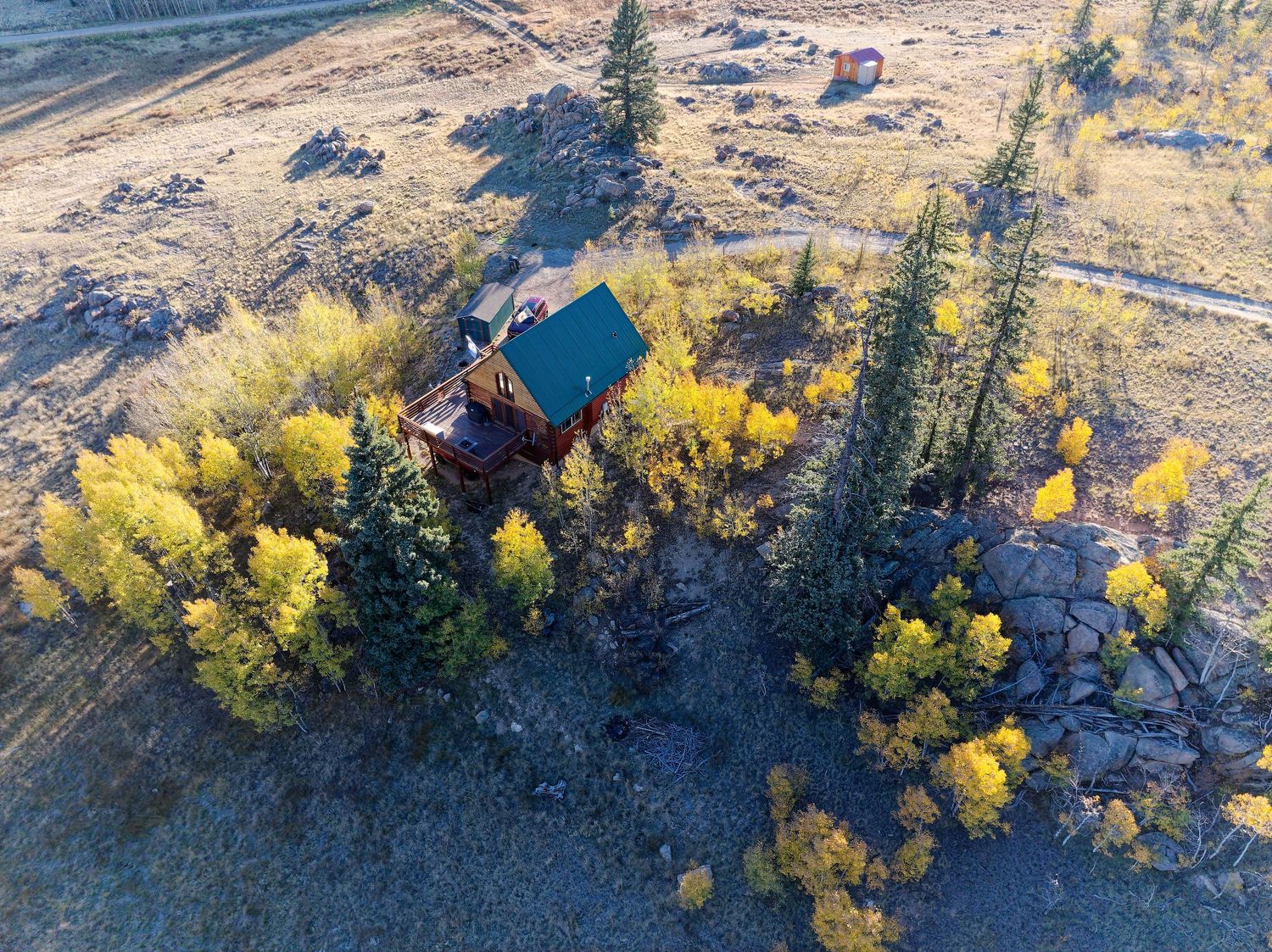 Aerial view of a rustic log cabin with green roof nestled among golden autumn aspen trees in a mountain valley.