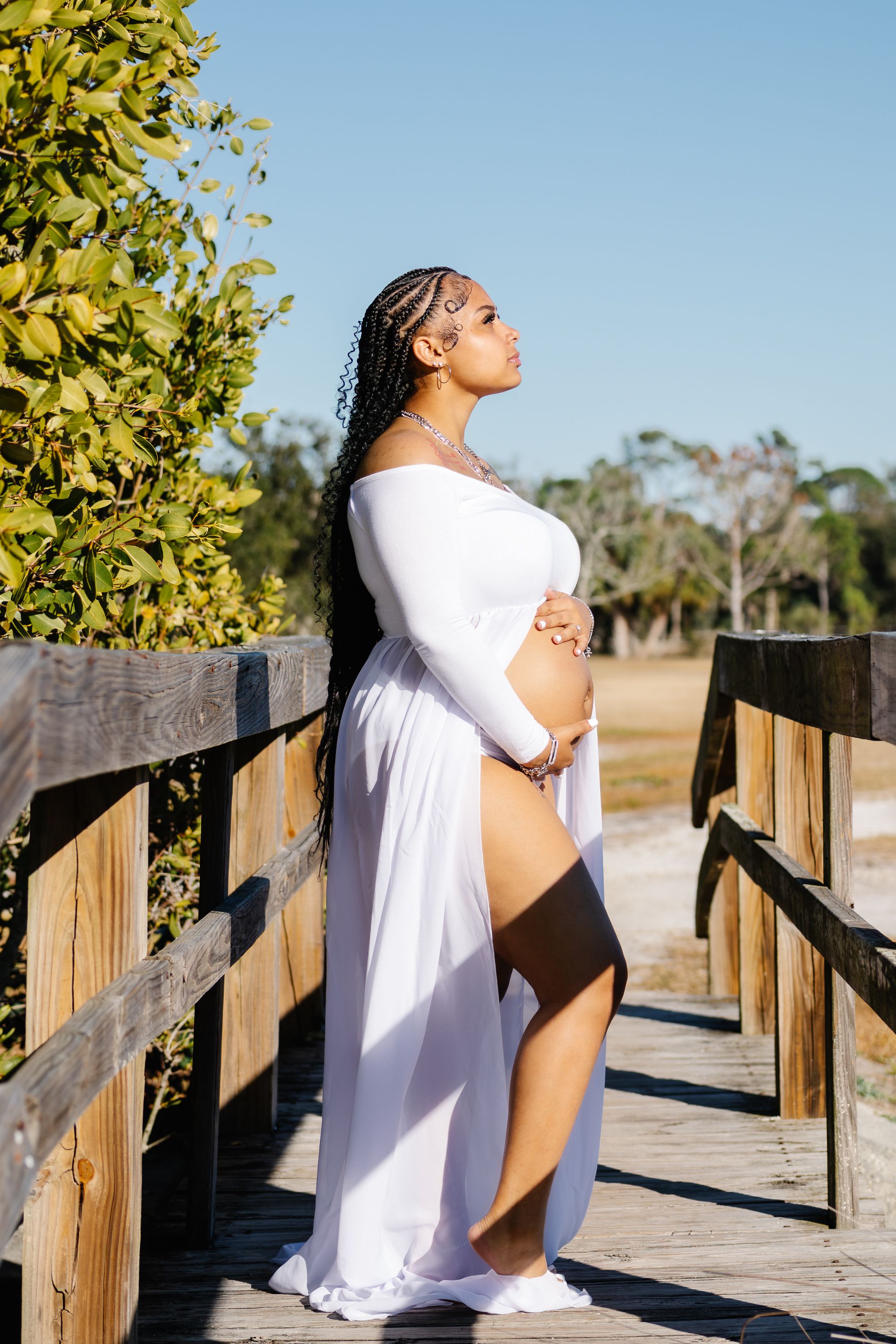 Pregnant woman in white off-shoulder gown cradling baby bump on wooden boardwalk surrounded by lush greenery.