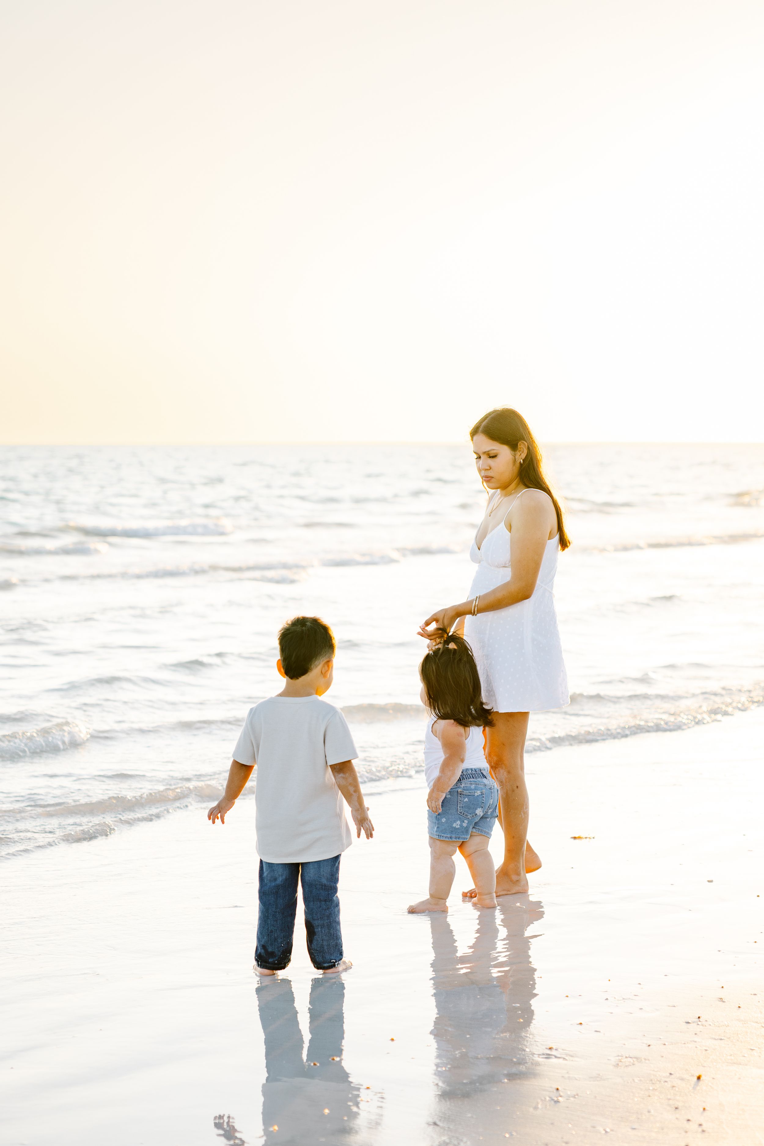 A mother and two young children playing at the ocean's edge during golden hour sunset on a sandy beach.