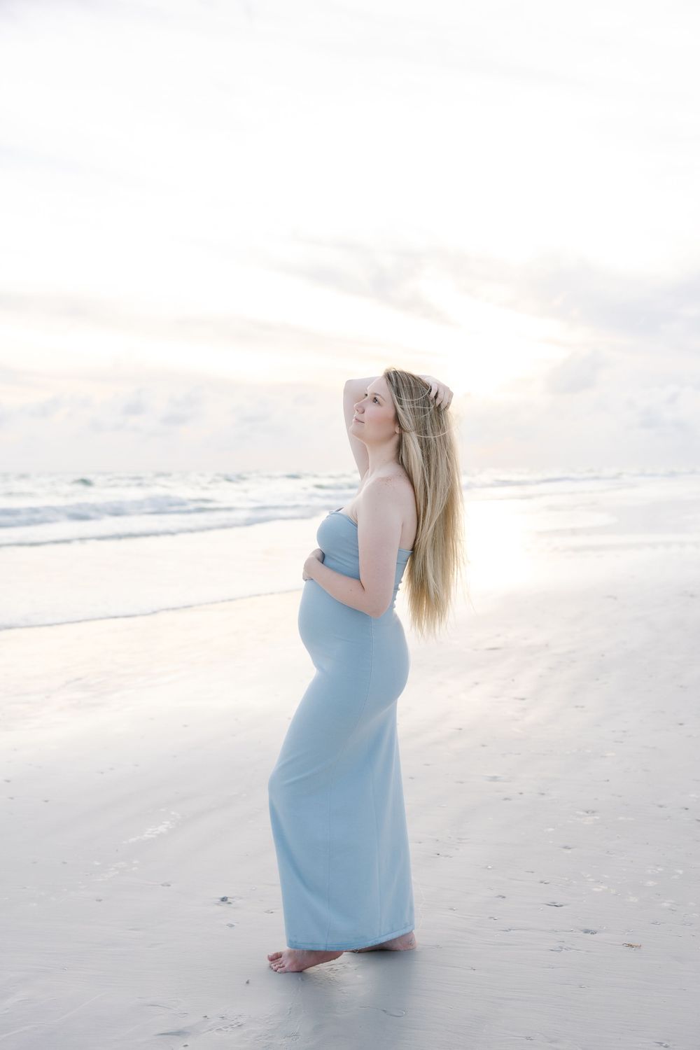 Pregnant woman in light blue maxi dress standing on beach at sunset, cradling baby bump with long blonde hair.