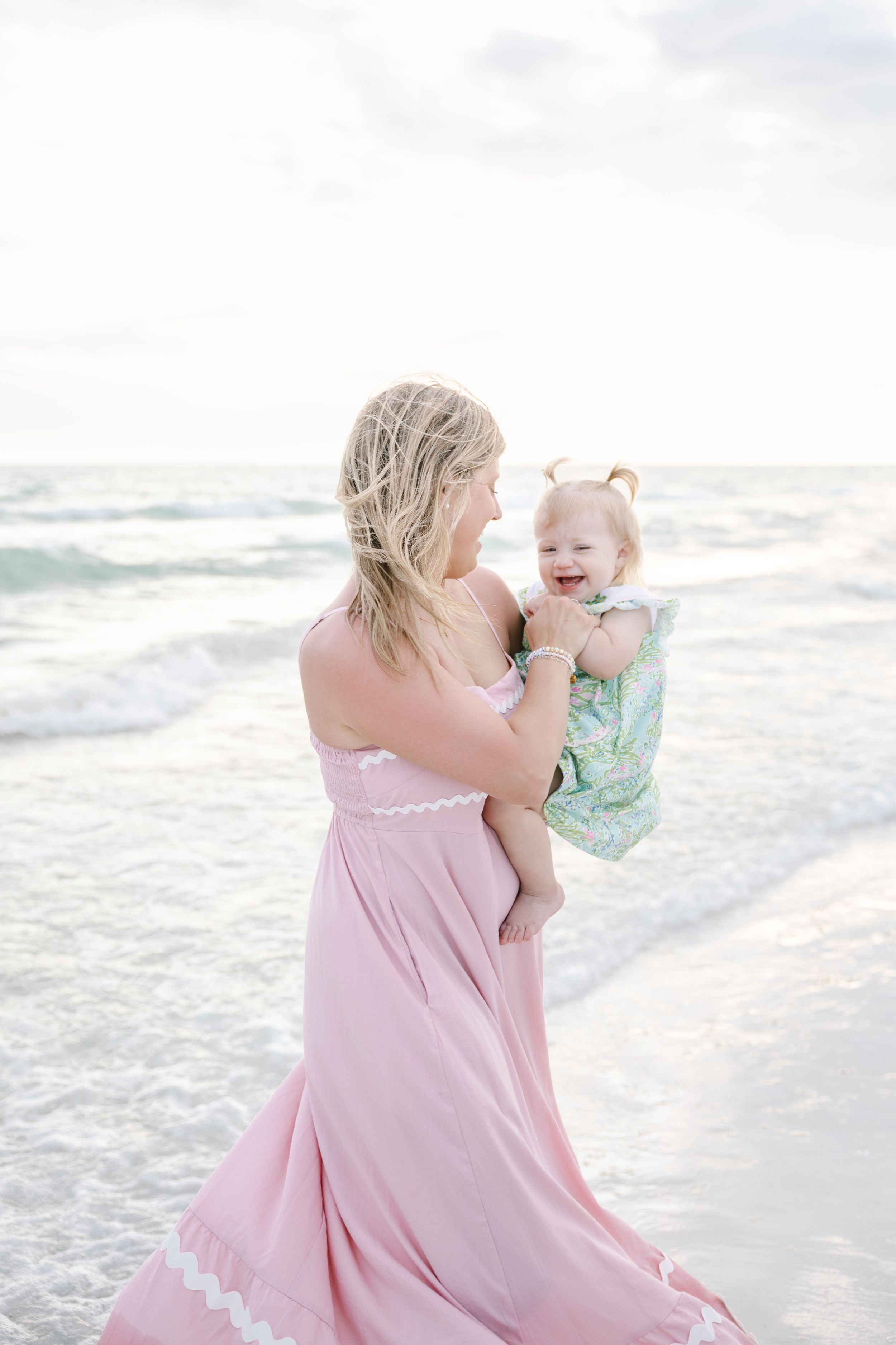 A mother in a pink dress holds her laughing baby girl on a beach with ocean waves behind them.