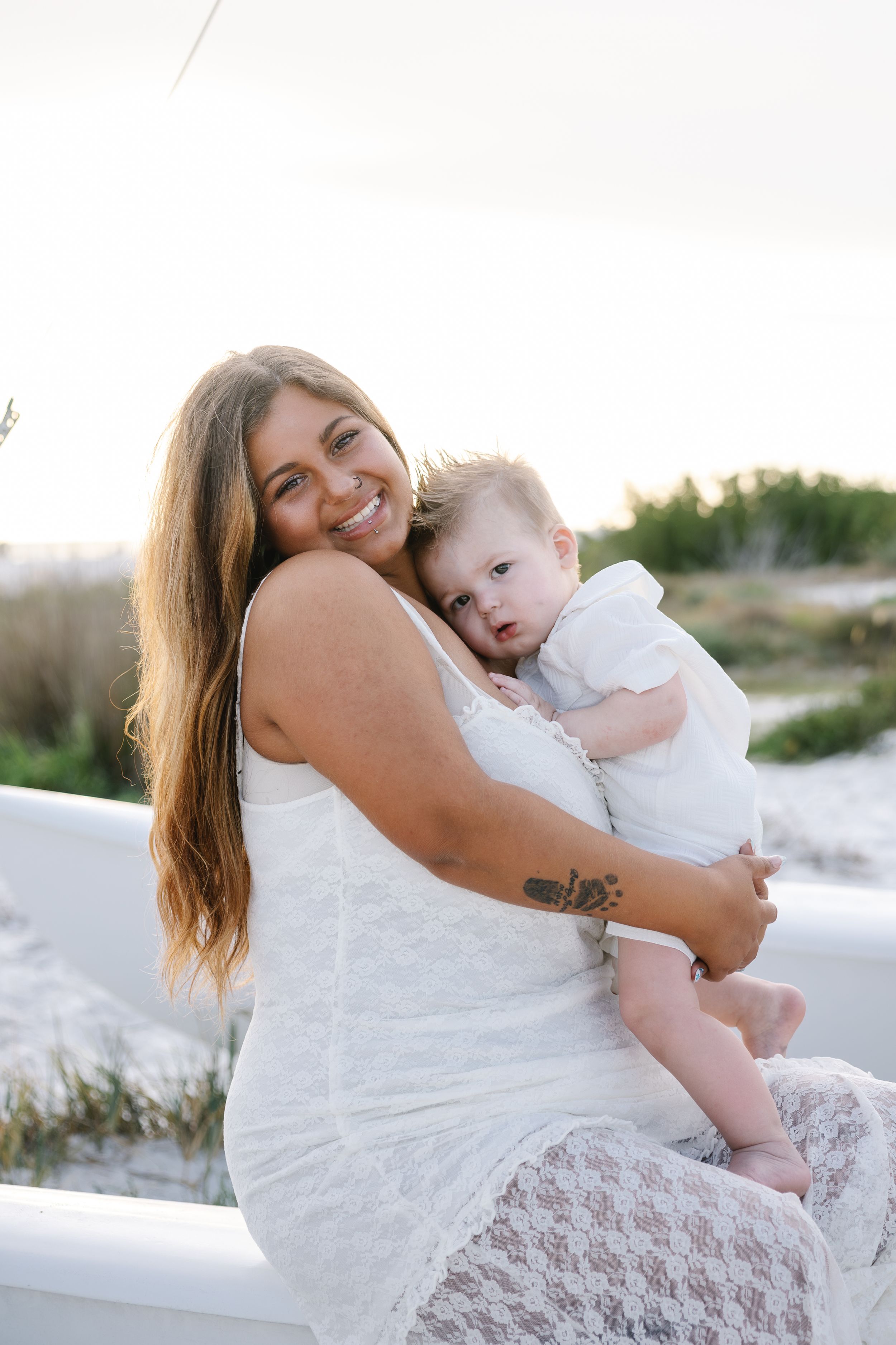 Smiling woman in white lace dress holding a baby in white outfit, sitting outdoors near sandy dunes at sunset.