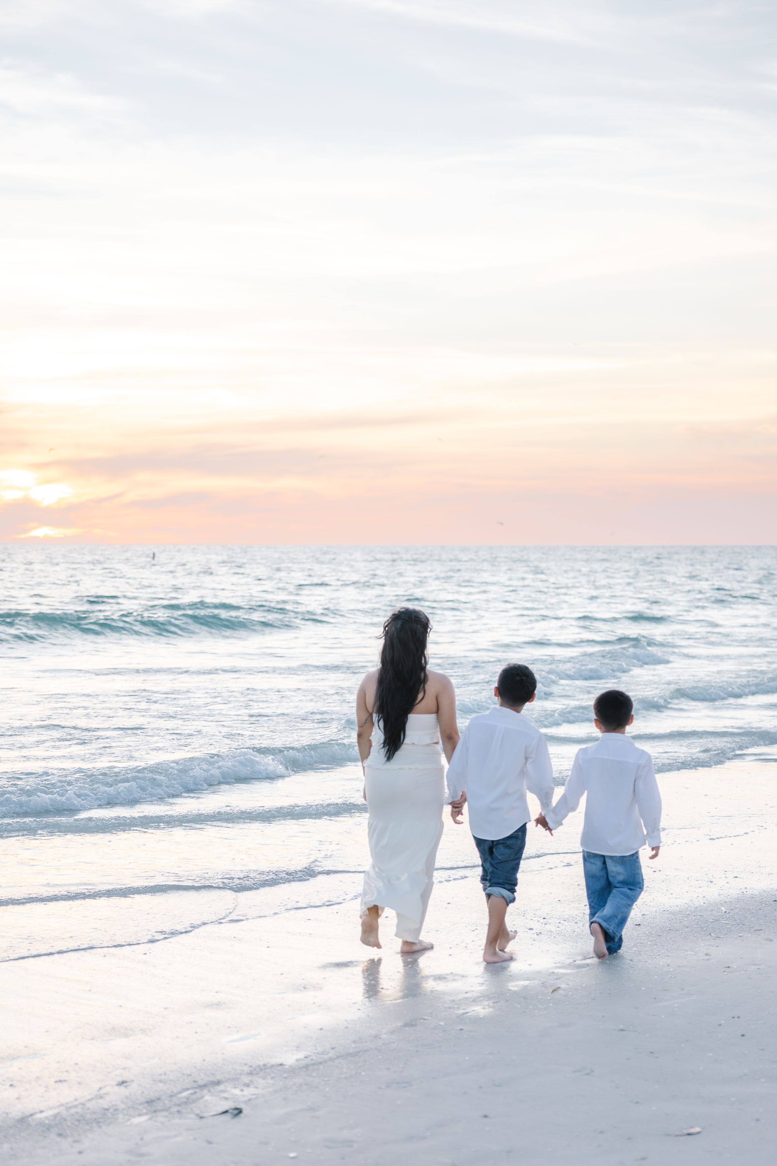 A mother and two children walk hand-in-hand along a sandy beach at sunset, dressed in white.