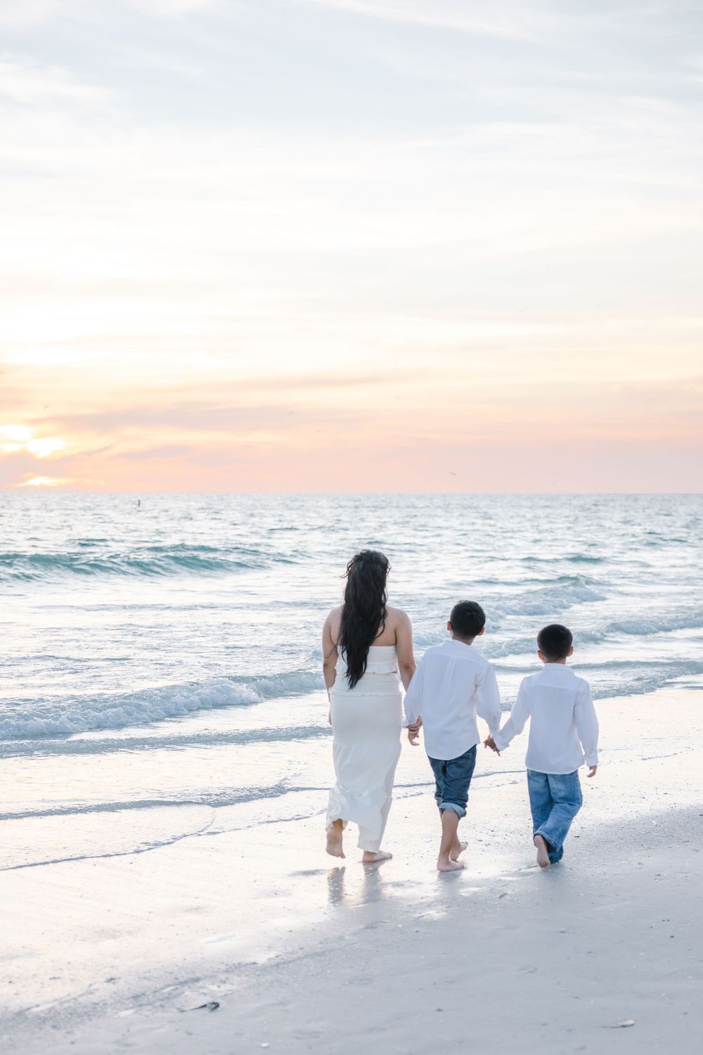 A mother and two children walk hand-in-hand along a sandy beach at sunset, dressed in white.