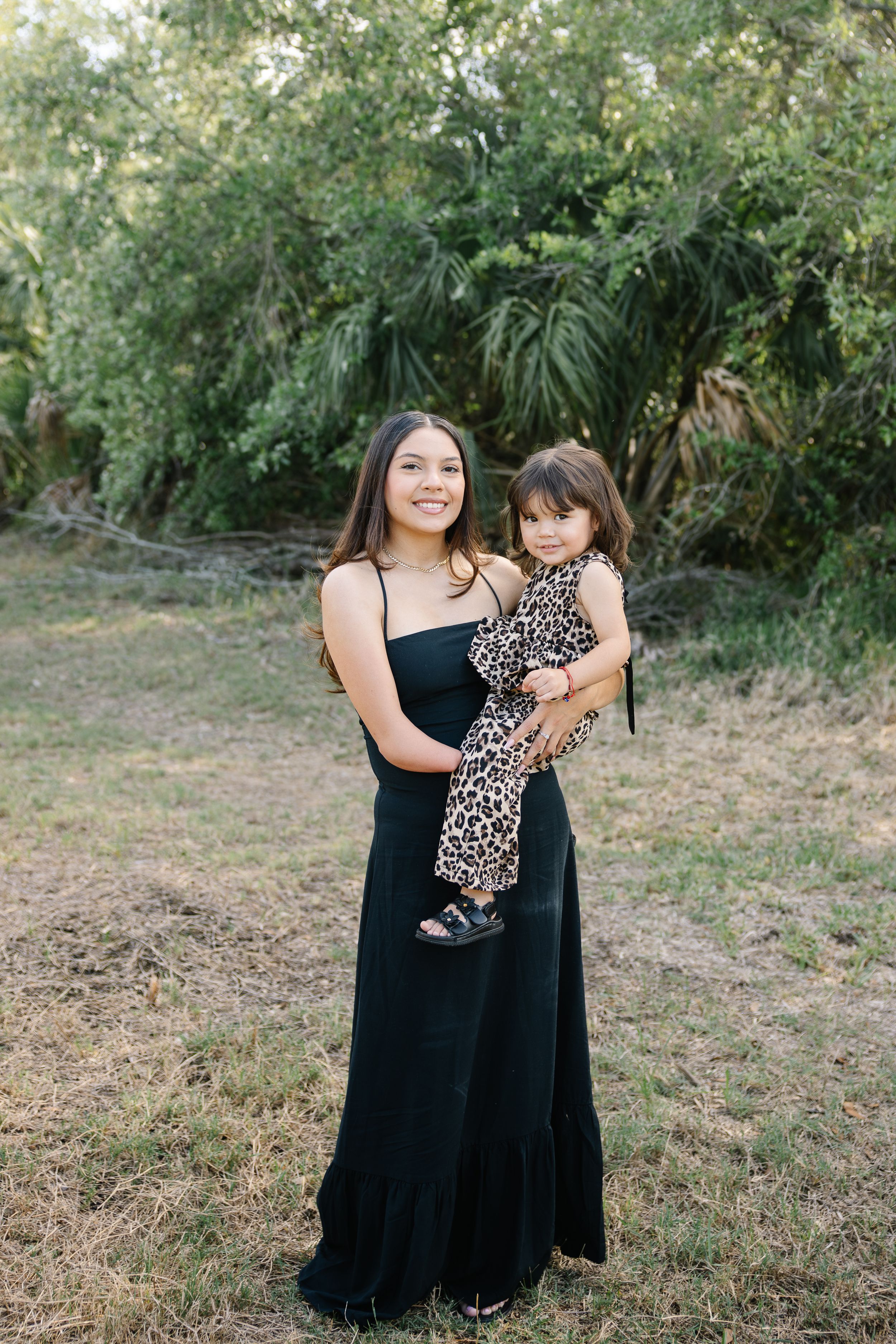 Young woman in black maxi dress holding a toddler in leopard print outfit outdoors near green trees.