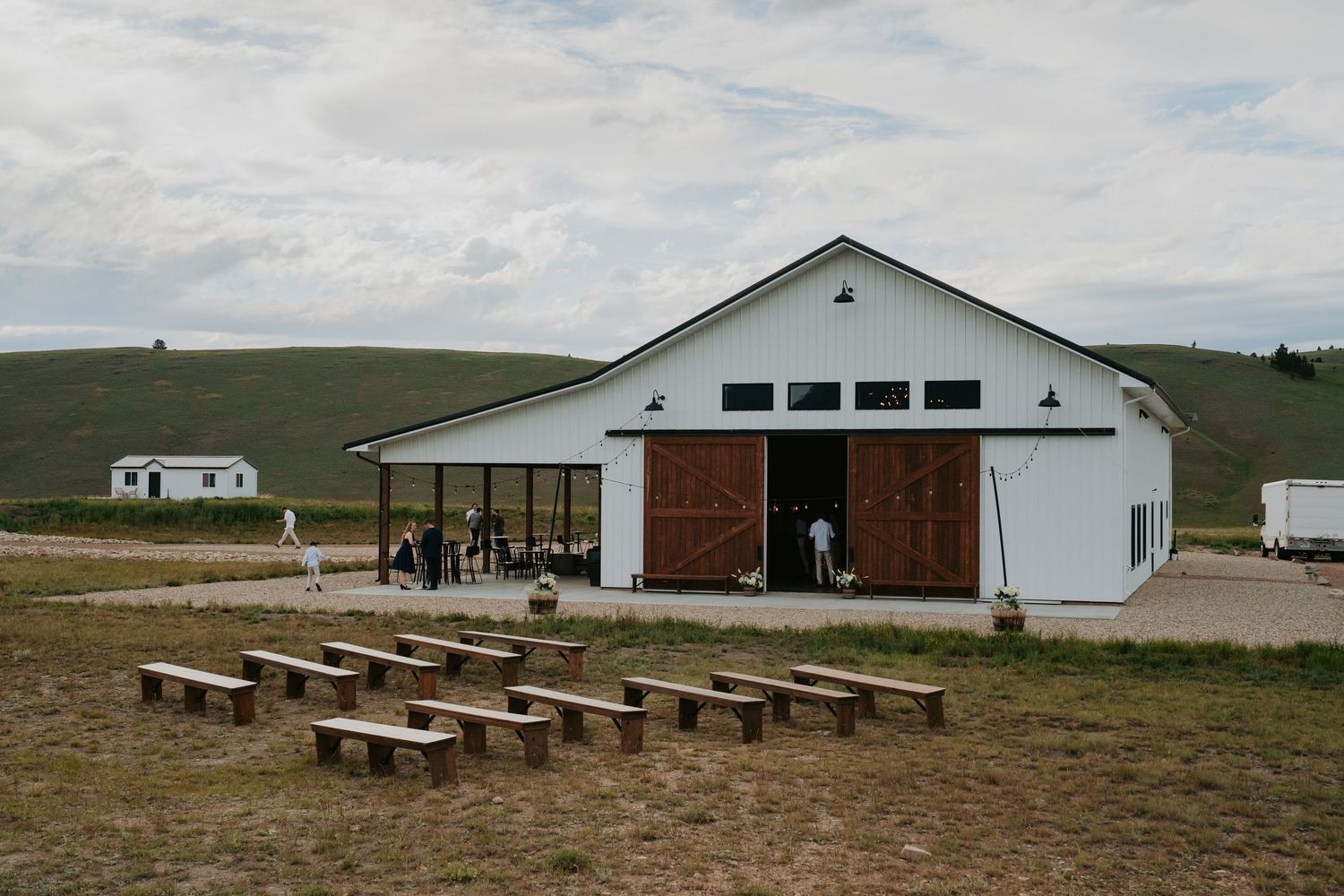 White barn venue with wooden doors, outdoor benches on dirt ground, rolling green hills backdrop, people gathering outside.