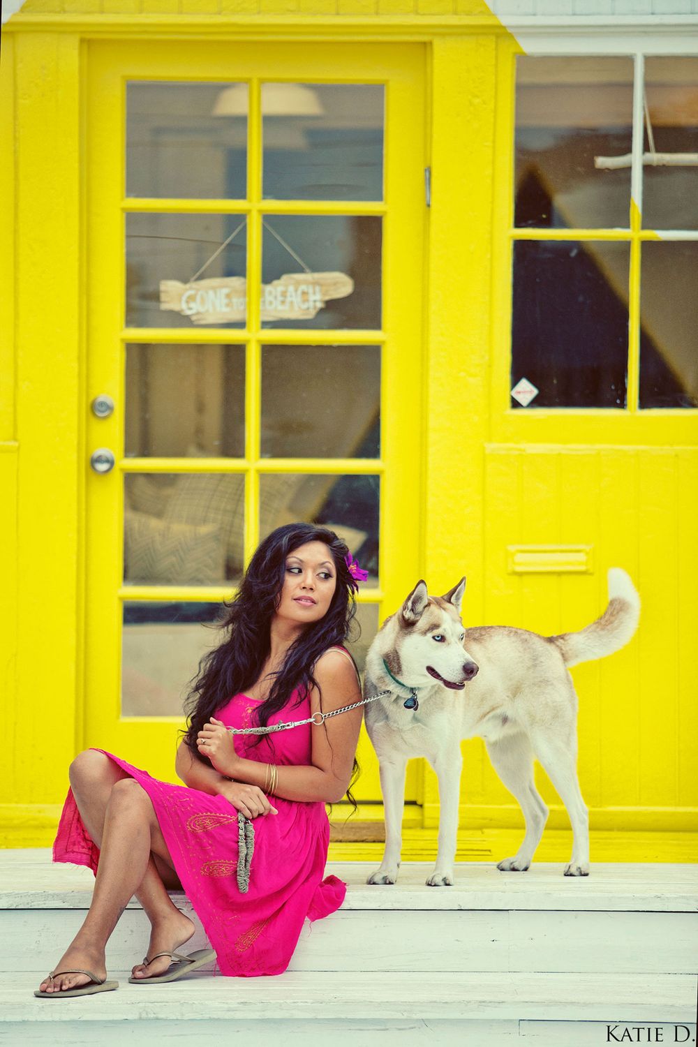 Woman in pink dress sits with a Husky dog in front of a bright yellow building exterior.