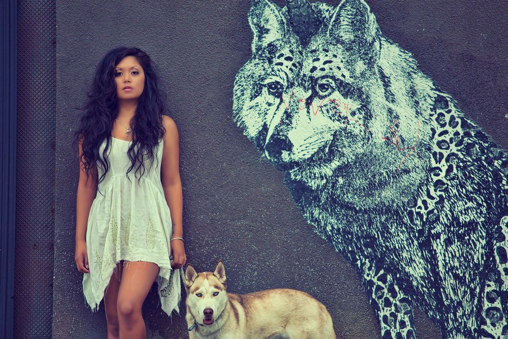 Young woman in white dress poses with a Husky dog beside a wolf street mural.