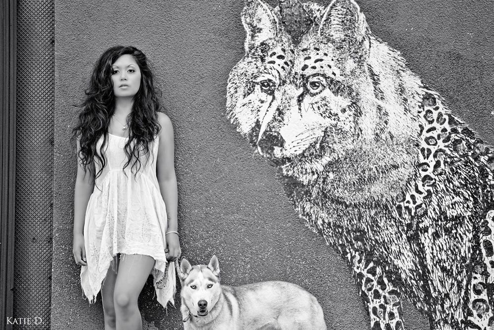 Black and white photo of woman with Husky dog beside a large wolf street art mural.