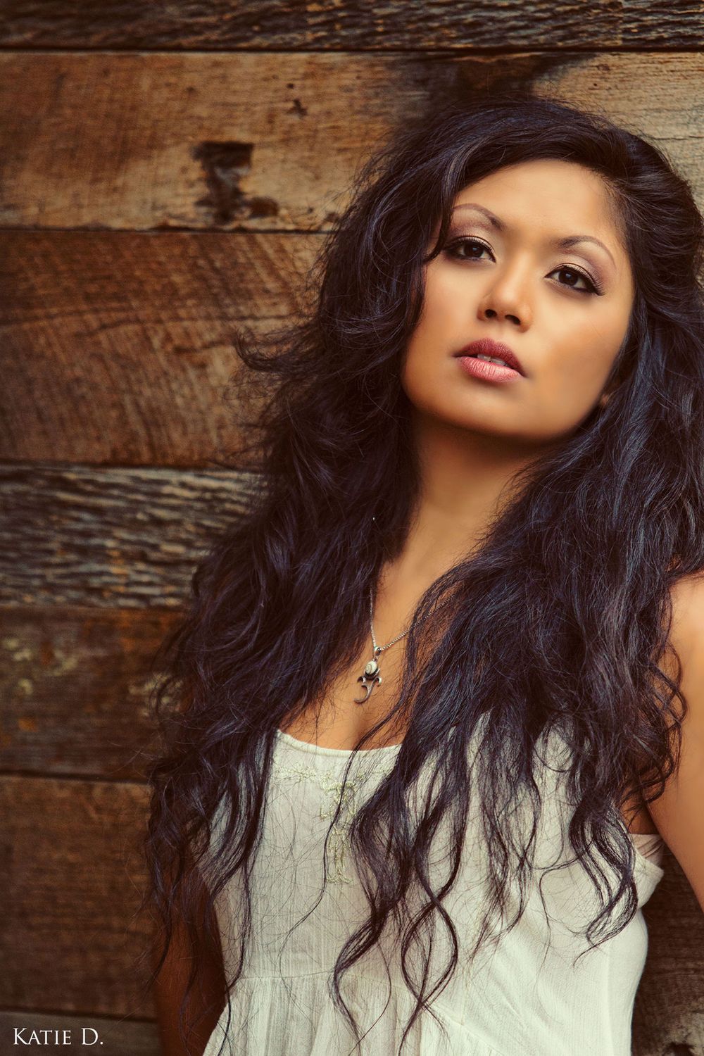 Beautiful woman with long dark wavy hair poses against a rustic wooden plank wall.