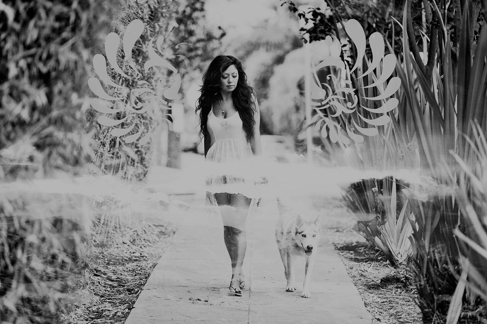 Black and white photo of a young woman in a white dress walking a husky dog on a garden pathway.