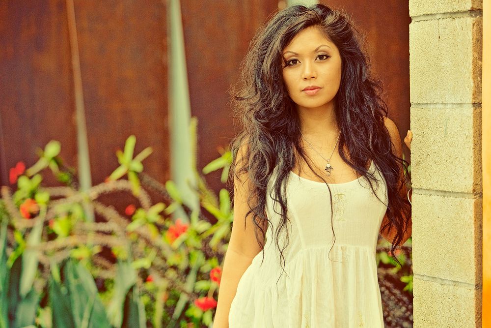 Young woman with long curly dark hair wearing a white sundress standing near a rustic wall.