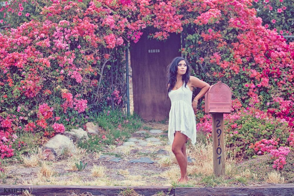 Young woman in a white dress standing by a mailbox surrounded by blooming pink bougainvillea.