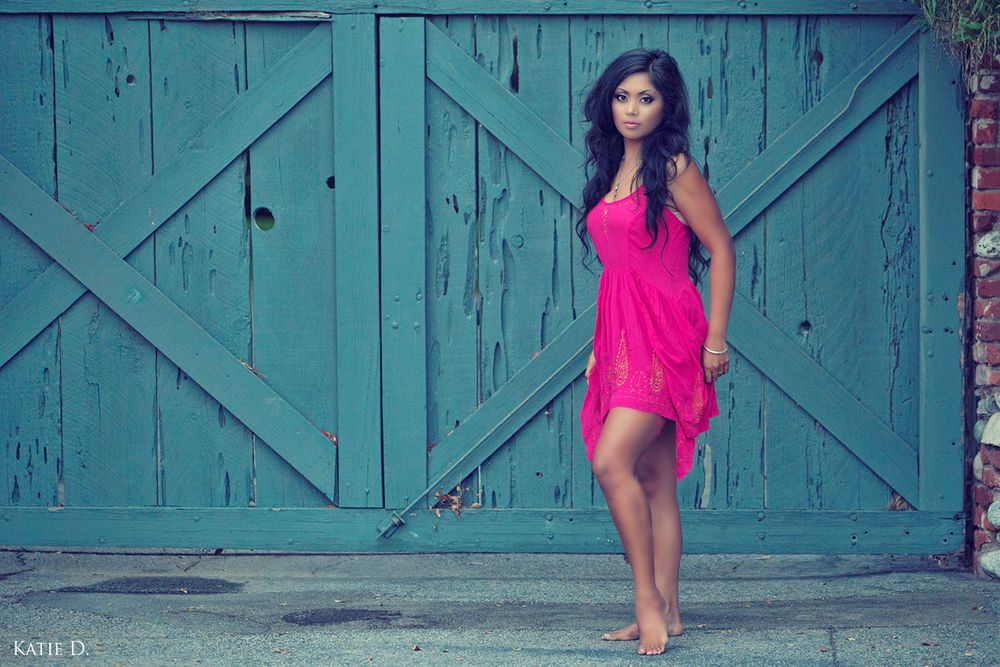 Young woman in a pink dress standing barefoot in front of rustic turquoise barn doors.