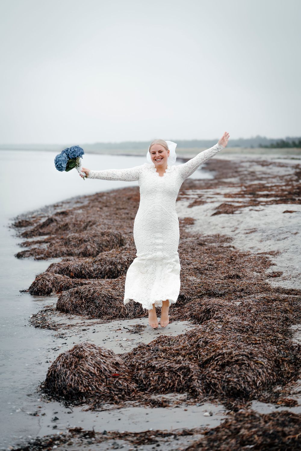 A person in a white dress stands joyfully on a seaweed-covered beach during a cloudy day.