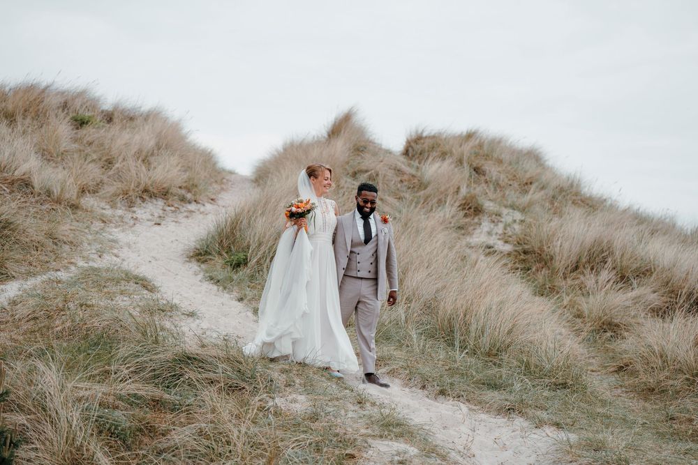 Bride and groom walking hand in hand through sandy dunes with tall grass on their wedding day.