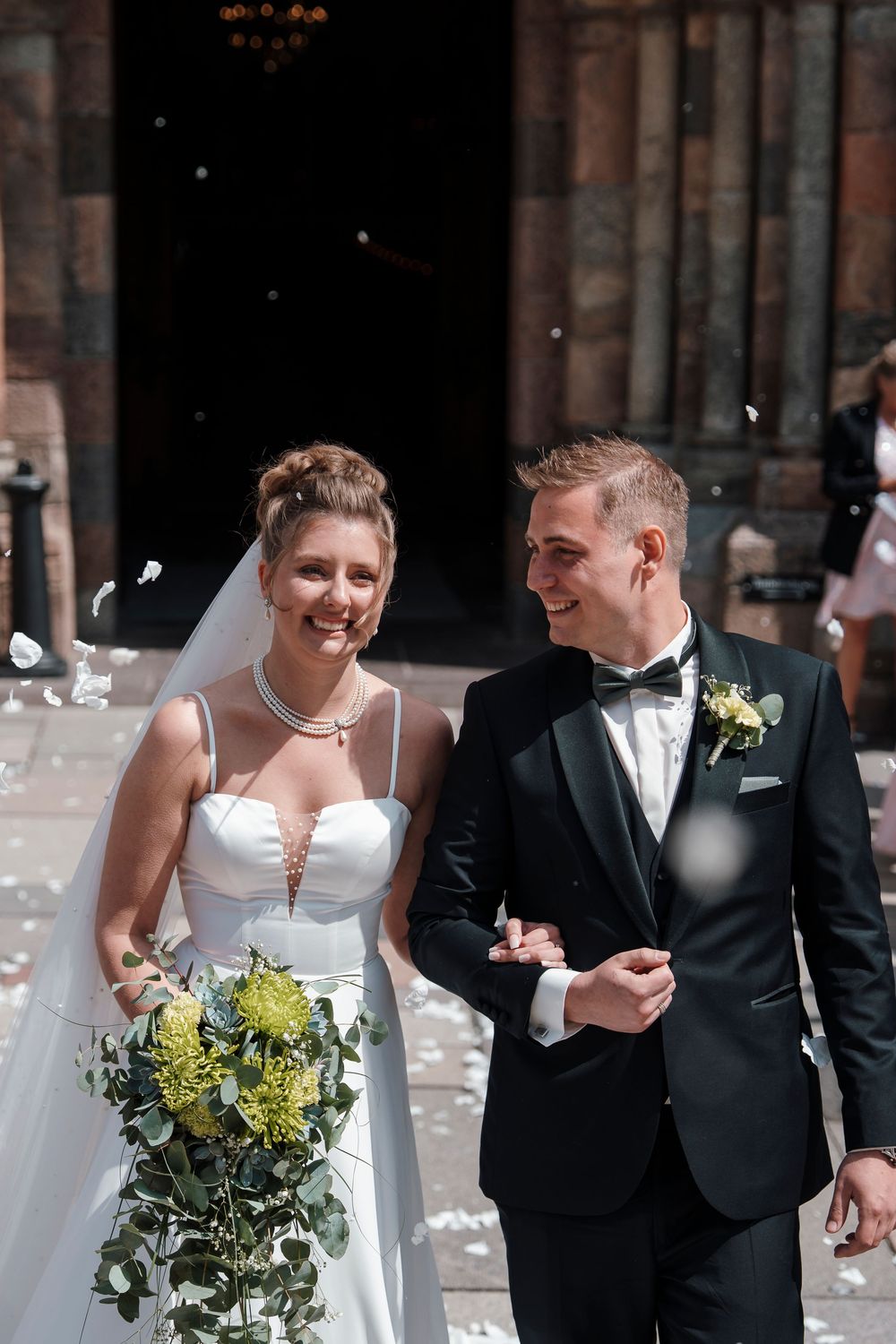 A couple in traditional wedding attire laughs joyfully together in front of rustic brick architecture.