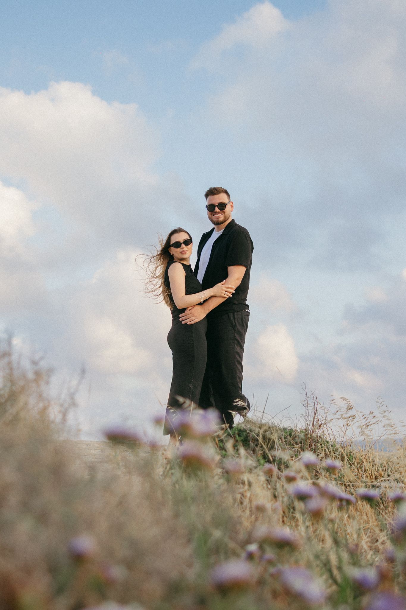 Couple in black outfits embracing on a hilltop with wildflowers and cloudy blue sky backdrop.