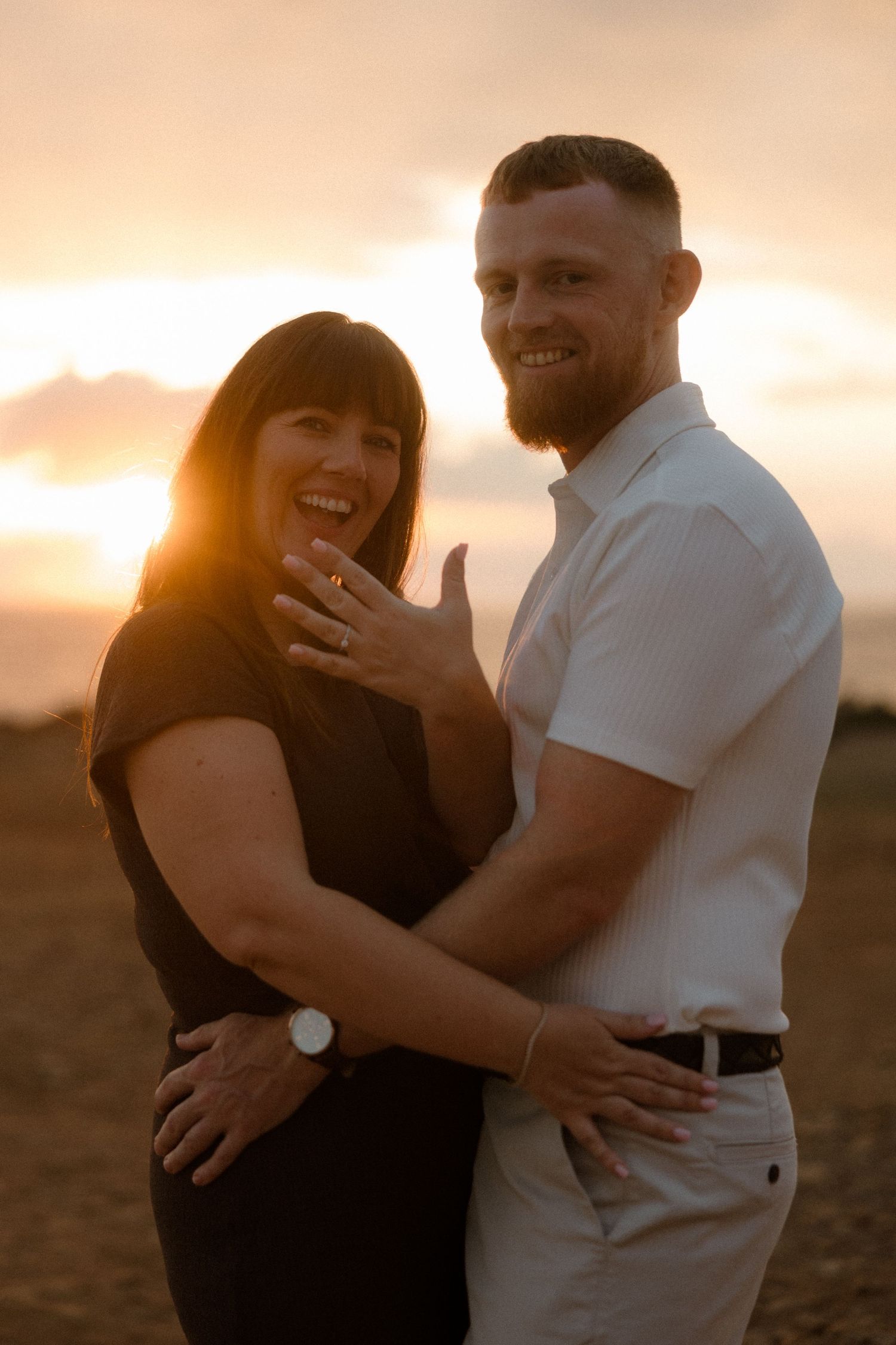 Couple in black outfits embracing on a hilltop with wildflowers and cloudy blue sky backdrop.