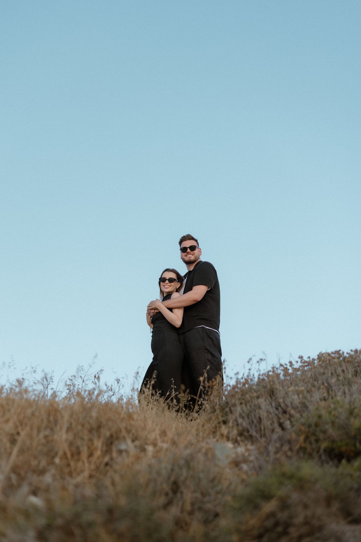 Couple in black outfits embracing on a hilltop with wildflowers and cloudy blue sky backdrop.