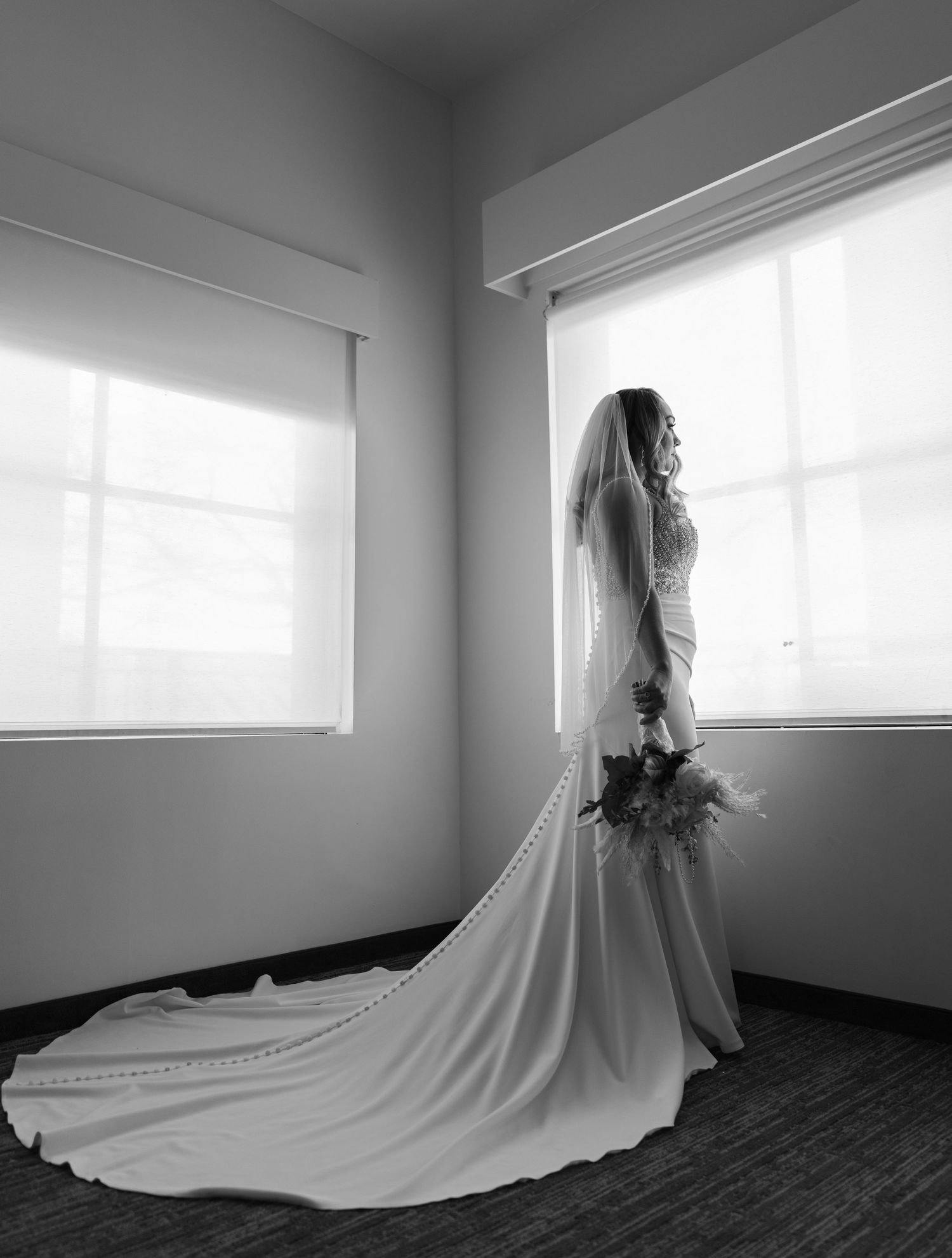 Black and white photo of a bride in a long gown and veil holding a bouquet, standing by bright windows.