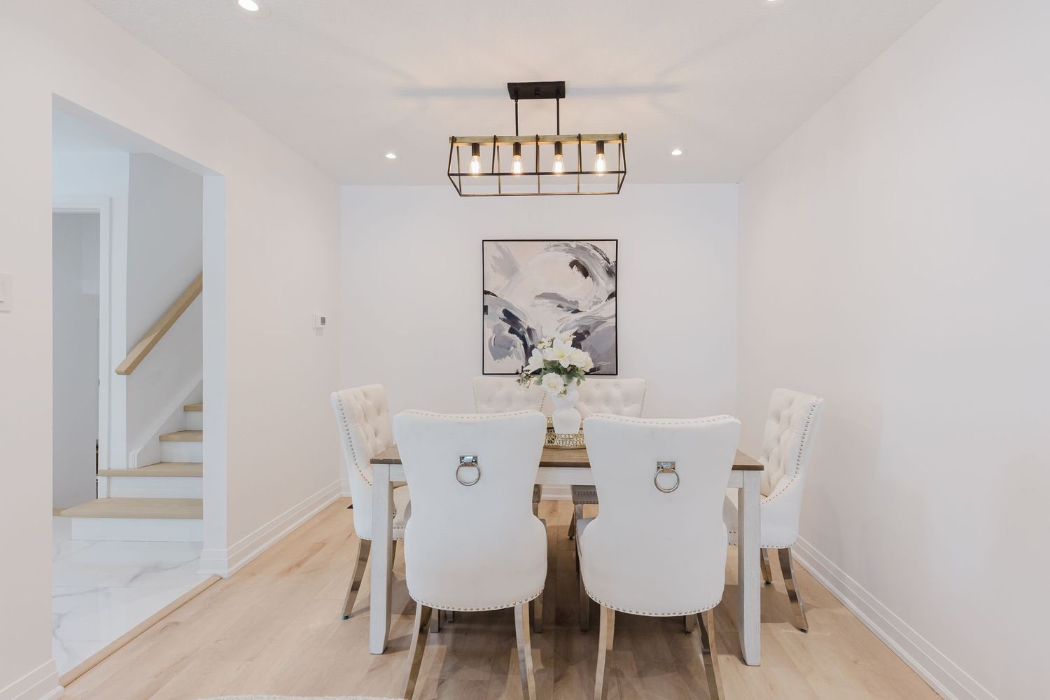 Elegant dining room with white tufted chairs, wooden table, modern chandelier, abstract wall art, and light hardwood floors.