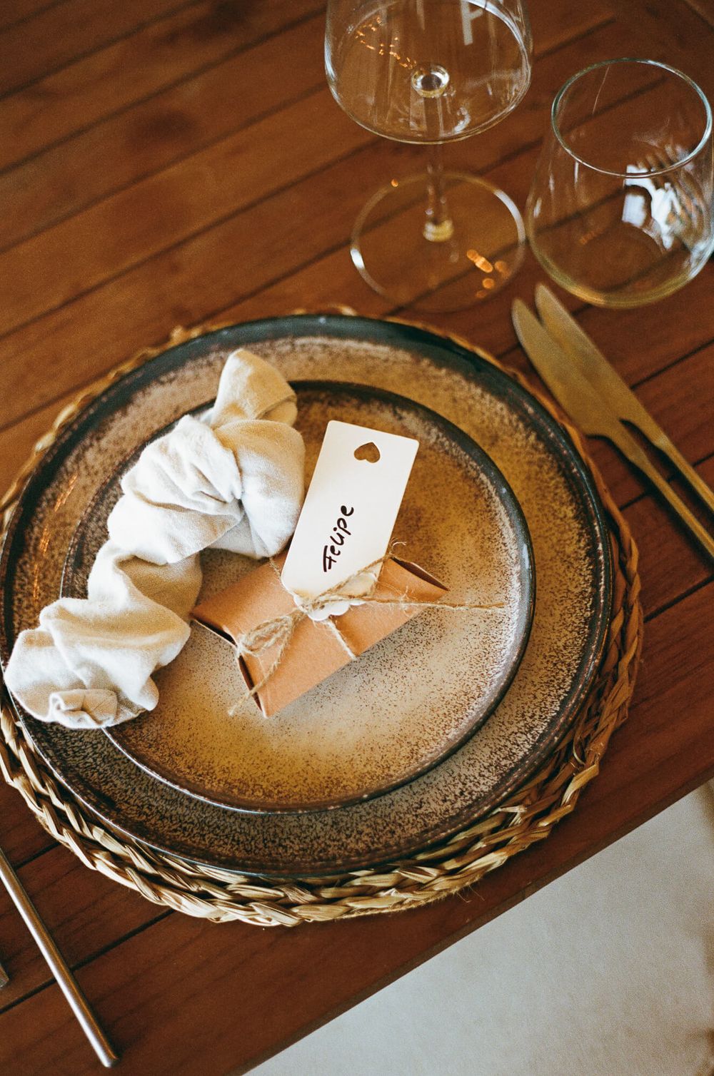 Elegant place setting with a rustic charger plate, white napkin, kraft paper gift, place card, and gold cutlery on a wood table.