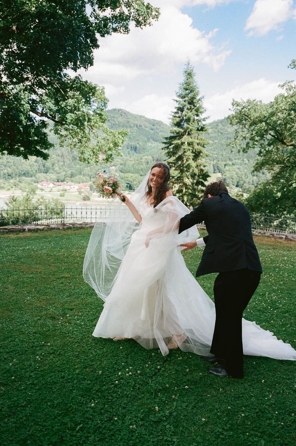 Bride in white tulle gown holds bouquet while groom adjusts her dress outdoors with green hills in background.