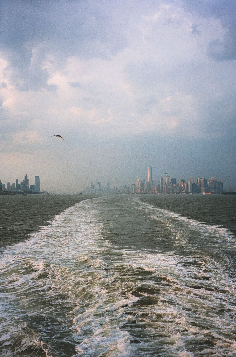 Boat wake on New York Harbor with Manhattan skyline and One World Trade Center visible under cloudy skies.