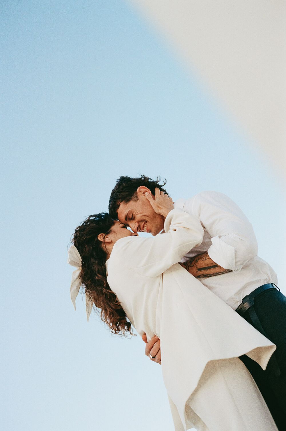 A couple shares a romantic embrace and kiss against a clear blue sky, the woman in a white dress with a bow.