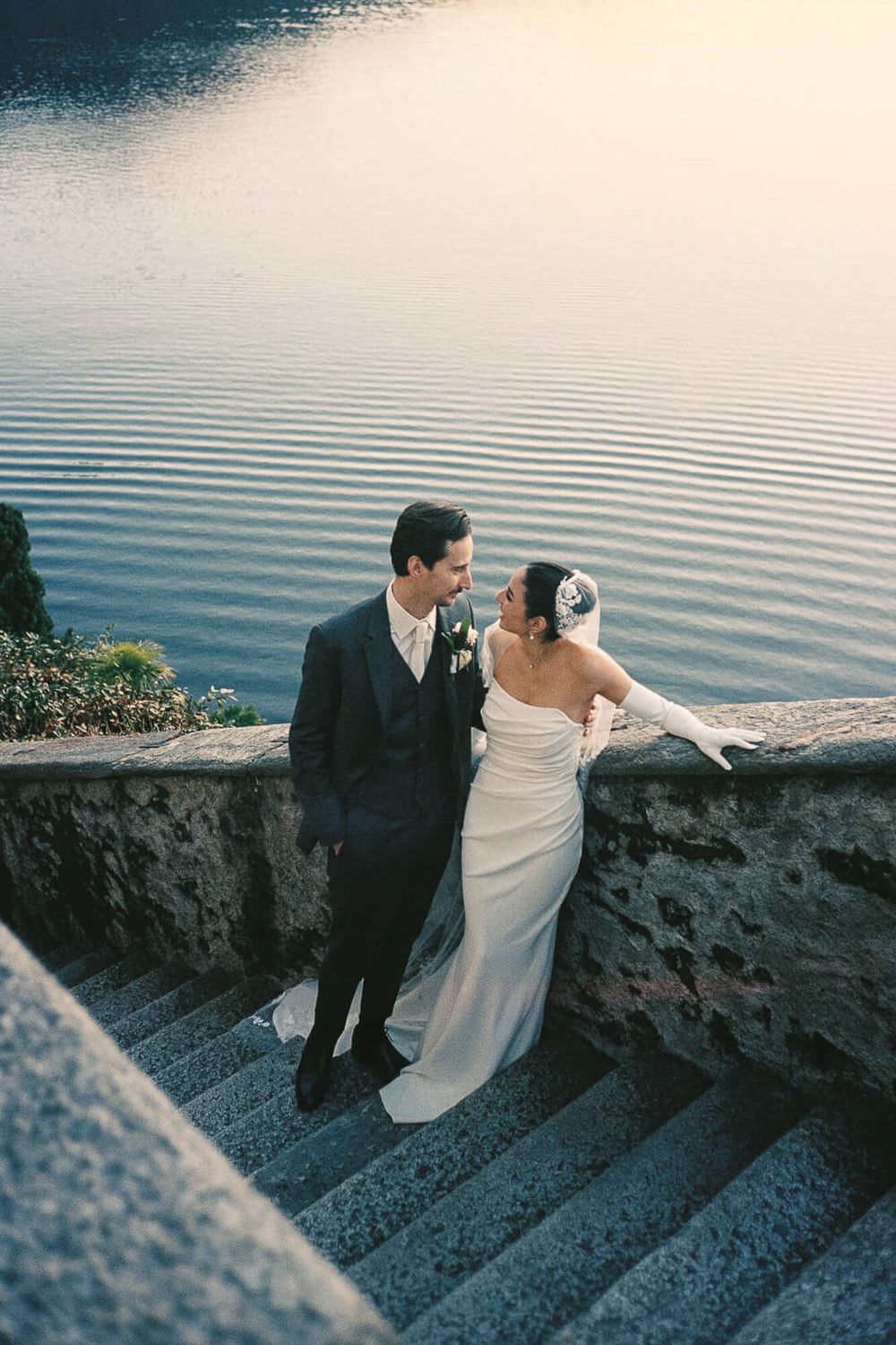 Bride and groom share a romantic moment on stone steps beside a serene lake at sunset on their wedding day.