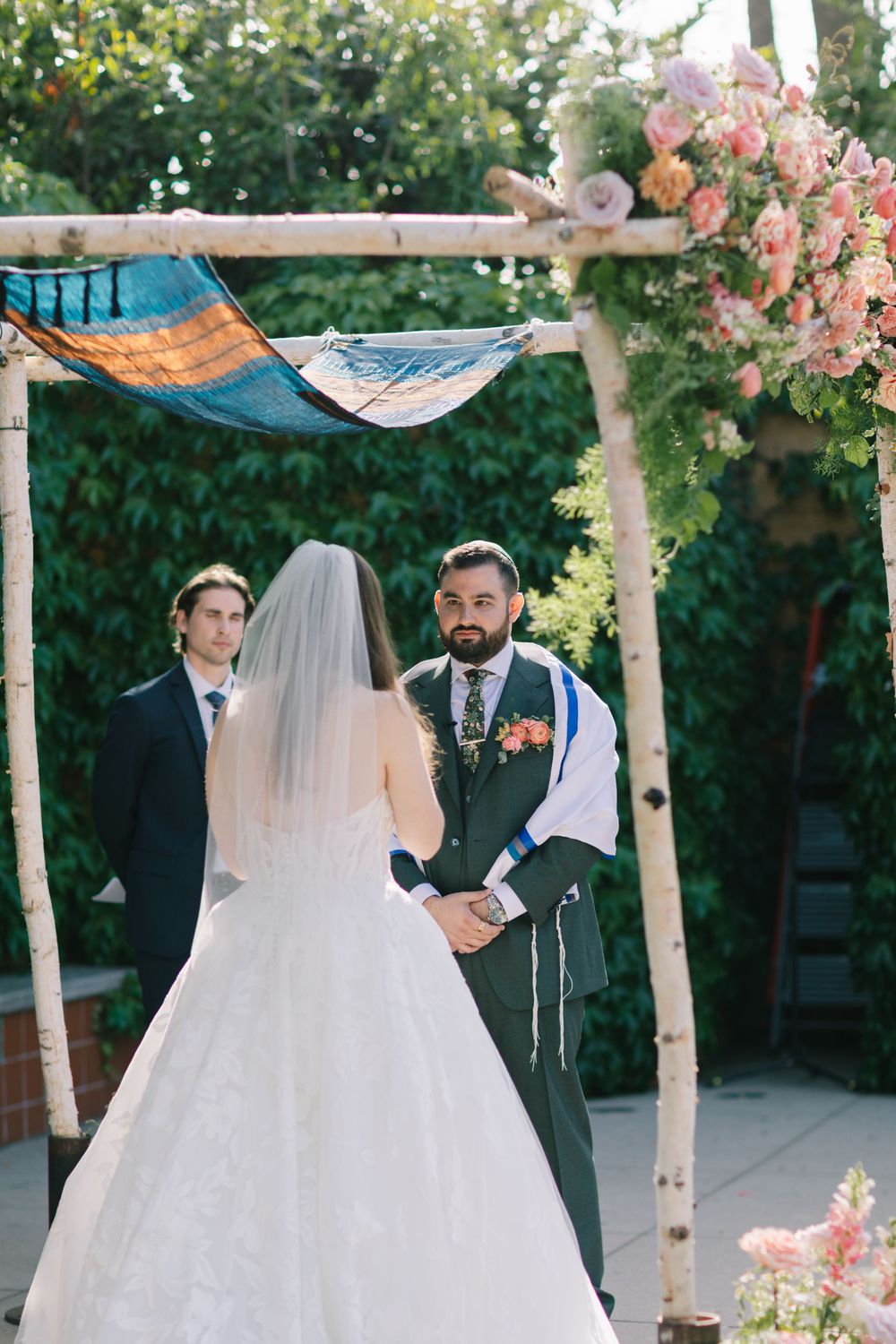 Groom gazing at bride during outdoor Jewish wedding ceremony under floral chuppah arch. The best California wedding photographer