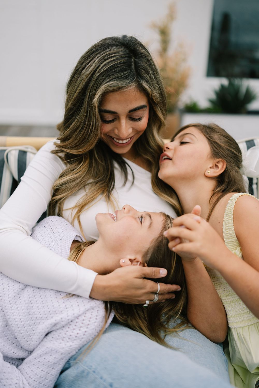 Laguna beach Family photography. Happy mother laughing and cuddling with two daughters on outdoor patio sofa together.