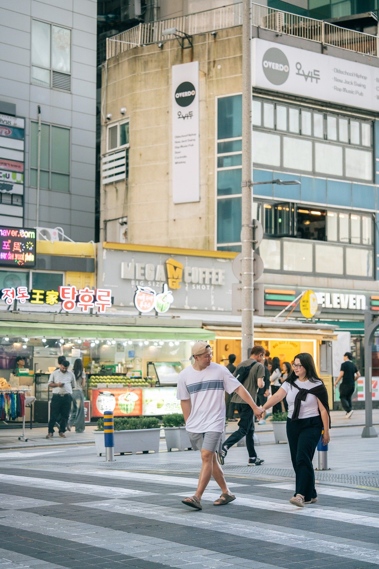 Couple holding hands crossing a busy urban street in Seoul, South Korea, with Mega Coffee and 7-Eleven stores visible.