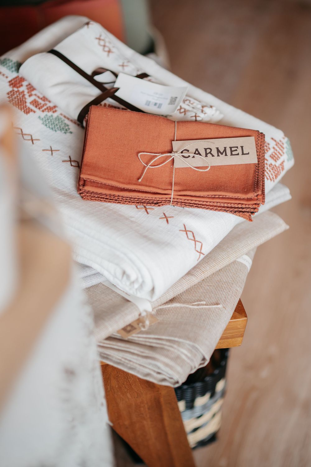 Folded terracotta fabric bundle labeled Carmel stacked on white embroidered textiles on a wooden shelf.