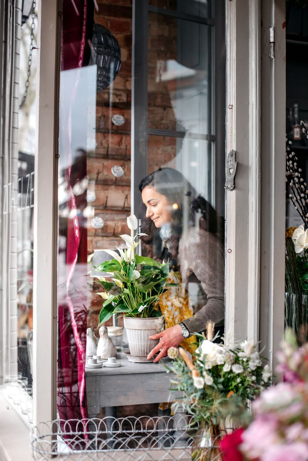 Smiling florist arranging white calla lilies in a flower shop window display with colorful blooms in foreground.