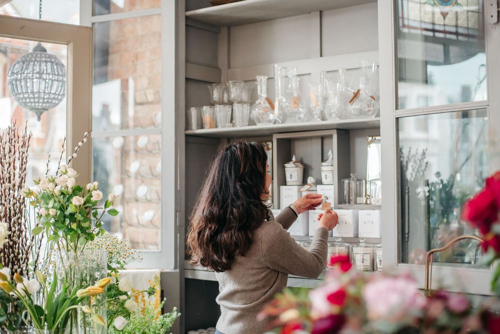 Woman arranging glassware on shelves in a bright floral shop surrounded by fresh flowers.