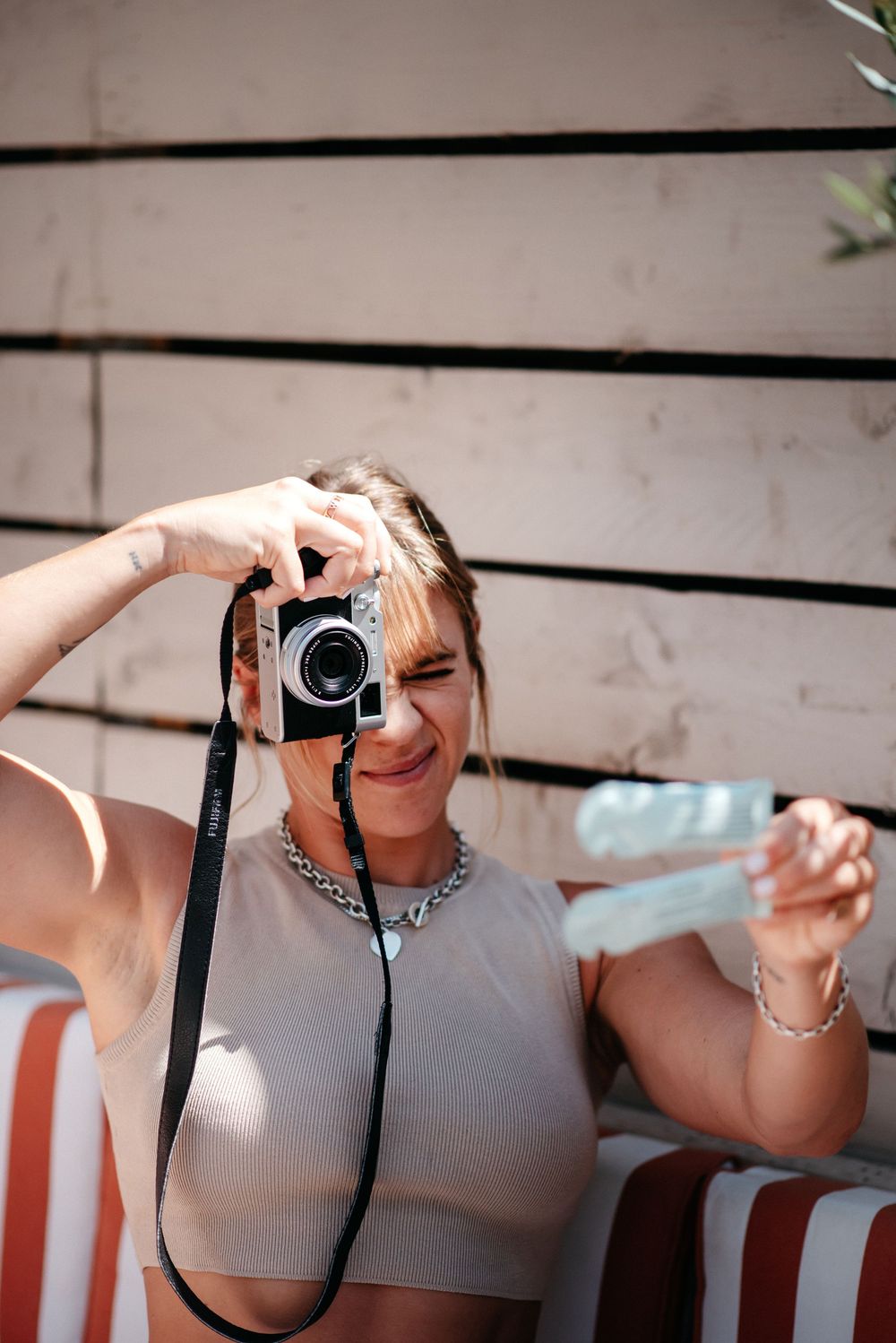 Young woman in beige crop top holding a film camera up to take a photo outdoors in bright sunlight.