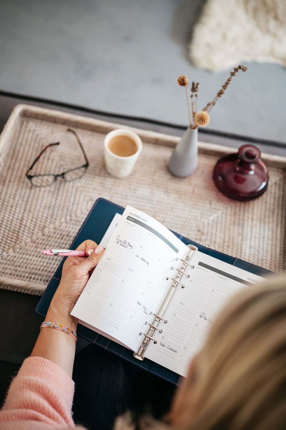 Woman writing in a ring-bound planner with a pencil, coffee cup, glasses, and small vases on a wicker tray nearby.