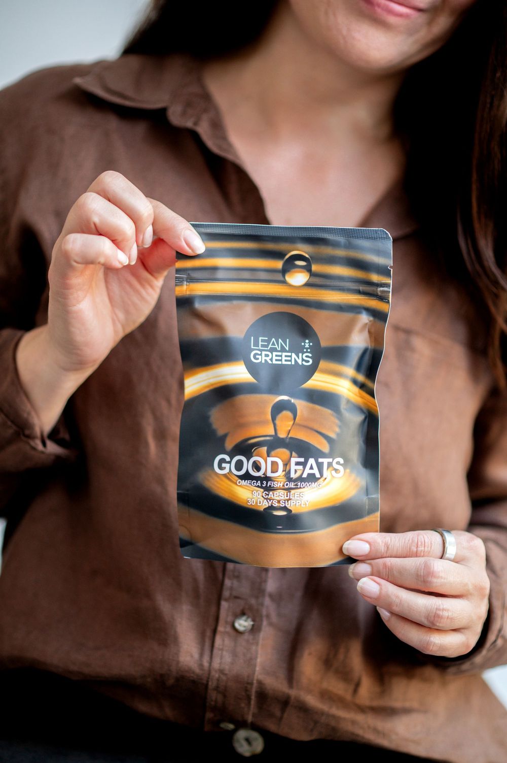 Woman holding a black pouch of Lean Greens Good Fats supplement powder while wearing a brown linen shirt.