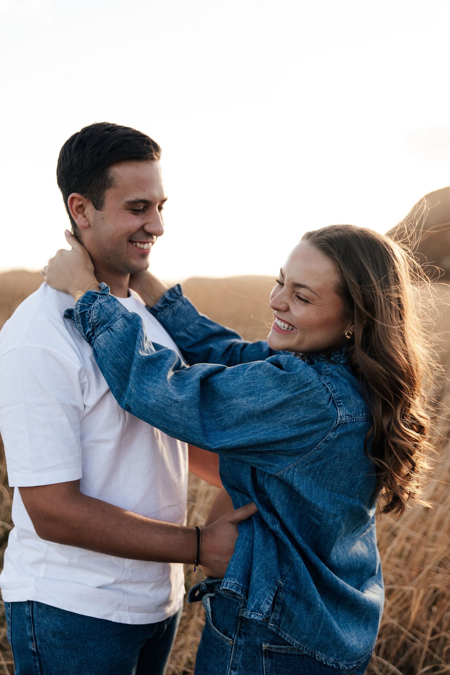 Happy couple embracing outdoors in golden field, woman in denim jacket laughing while holding man in white t-shirt.