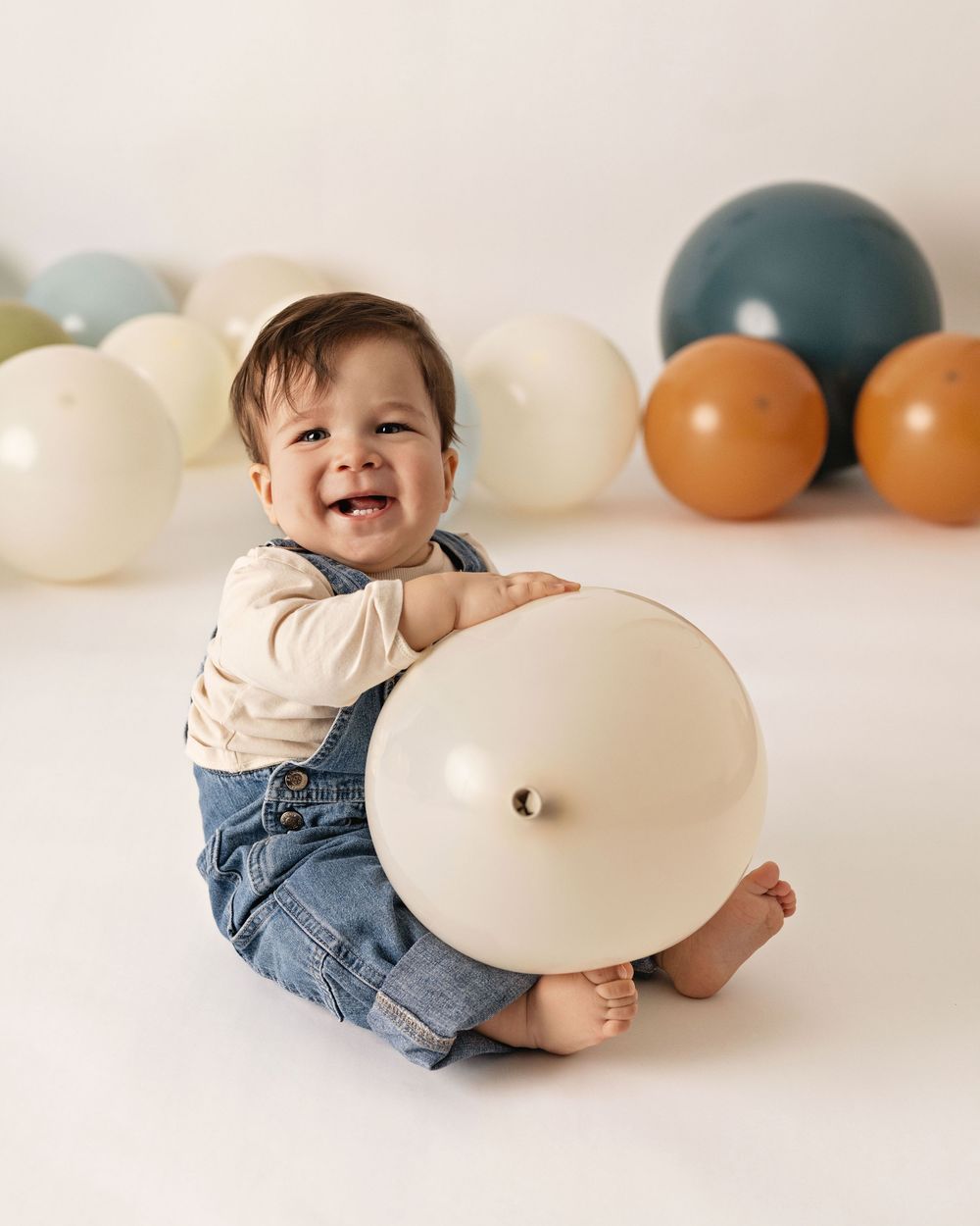 Smiling baby in denim overalls sitting on white floor hugging a large cream balloon with colorful balloons behind.