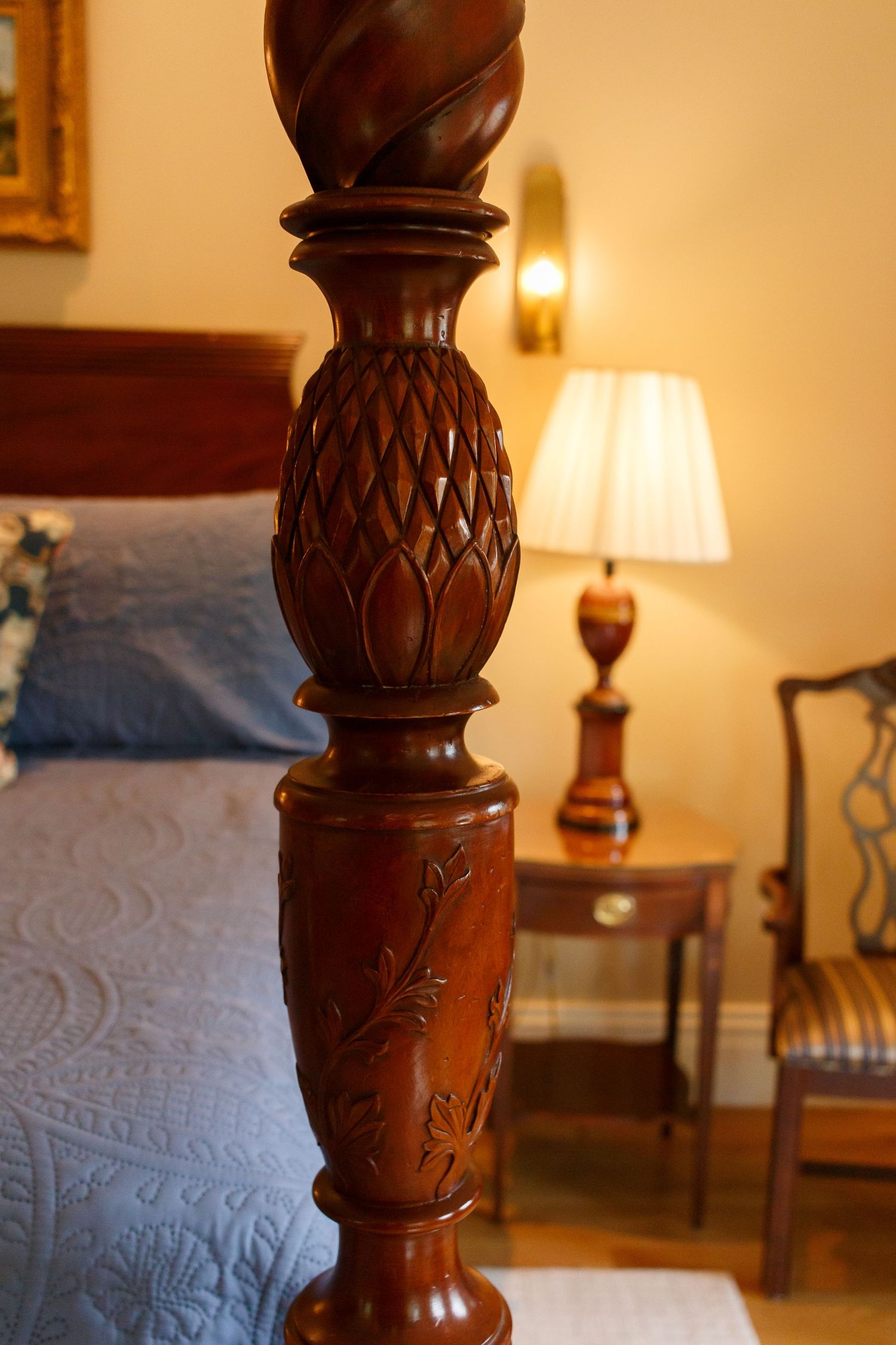 Ornately carved mahogany four-poster bed post with pineapple detail in an elegant bedroom with warm lamp lighting.