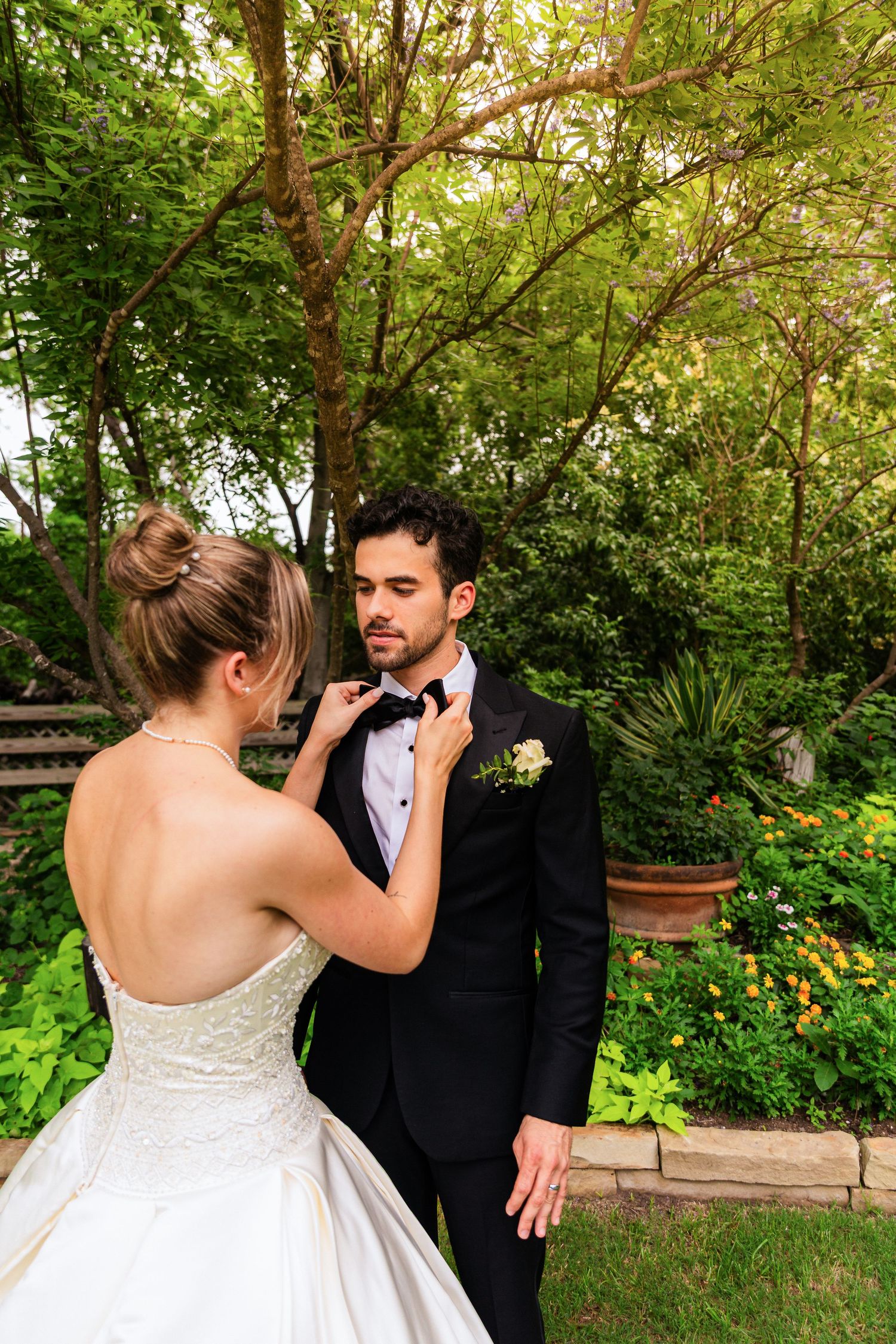 Bride in strapless lace gown adjusting groom's bow tie in a lush garden setting on their wedding day.