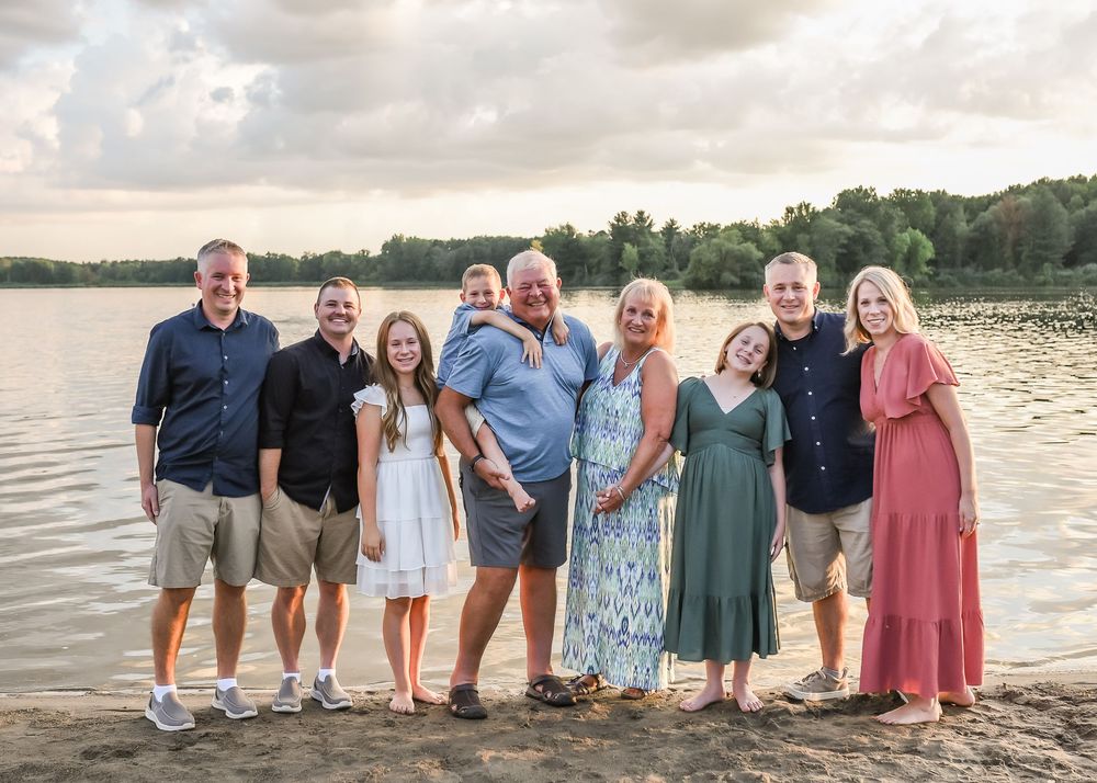 Family of nine posing together on a lakeside beach at sunset with lush green trees in the background.