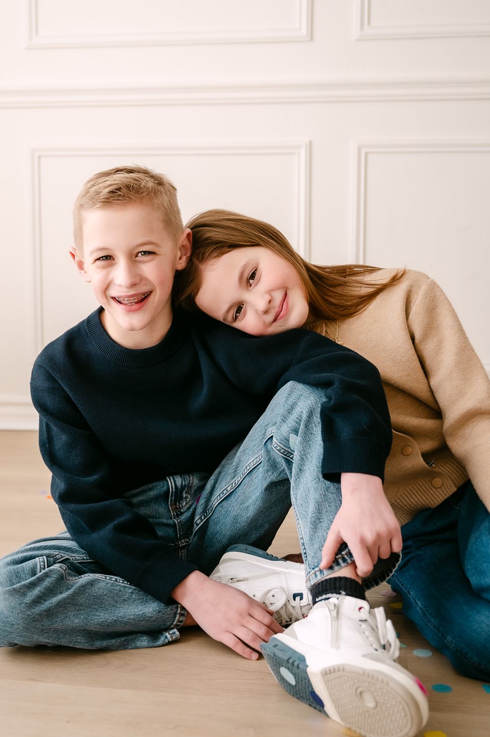 Two smiling children sit on floor, girl in tan sweater leans on boy in navy sweatshirt, both wearing jeans.